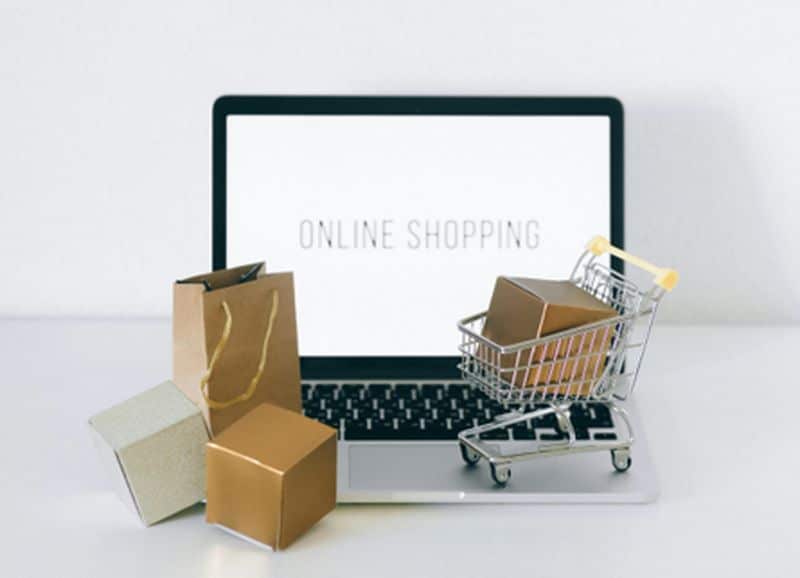 מחשב נייד המציג "קניות אונליין" מציג לראווה עגלת קניות מיניאטורית עם קופסה קטנה על המקלדת, מוקפת בכמה קופסאות קטנות ושקית קניות מנייר, הממחישה את המהות של מסחר אלקטרוני.