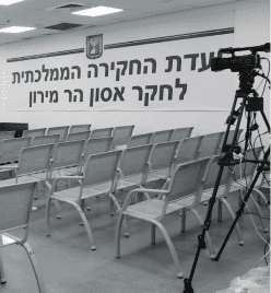 תמונה בשחור-לבן מתארת חדר ריק, שבו שורות של כיסאות מתכת פונות לקיר המעוטר בטקסט עברי גדול. בצד ימין, מצלמת וידאו עומדת על חצובה. אווירה של ציפייה ממלאת את המסגרת הזו של חבר תרגומים.