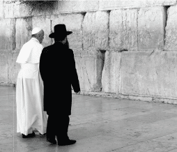 שני מנהיגים דתיים - האחד בחלוק וכובע לבן, השני במעיל שחור וכובע רחב שוליים - עומדים זה לצד זה מול קיר אבן גדול. הסצנה, שצולמה בשחור-לבן, מעוררת אחווה שקטה המזכירה חבר תרגומים.