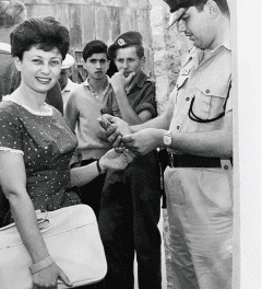 אישה מחייכת כשגבר במדי צבא בודק את המסמכים שלה, אולי דן בתפקידה החדש כחבר תרגומים. שני צעירים עומדים ברקע, מתבוננים במתרחש. נראה שהסביבה נמצאת בחוץ באזור עירוני.