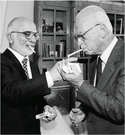 שני גברים מבוגרים בחליפות עומדים בחדר עם מדפי ספרים. שניהם מחזיקים סיגריות ומצית, מסייעים זה לזה בהדלקת הסיגריות שלהם. זה כאילו הם מעורבים בסוג של חבר תרגומים, מתרגמים את אמנות השיחה להבנה הדדית.