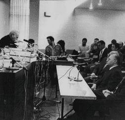 תמונה בשחור לבן מצלמת חדר ישיבות של חבר תרגומים. ההתקנה כוללת רמקול במיקרופון מוקף בציוד, מול קהל. חלק מהמשתתפים רושמים הערות בעוד אחרים מקשיבים בקשב רב בחלל המעוצב בפשטות.