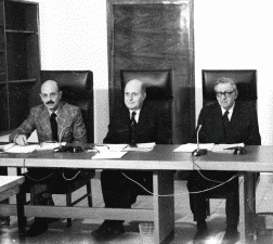 שלושה גברים בחליפות יושבים מאחורי שולחן גדול עם ניירות ומיקרופונים לפניהם, ודנים לכאורה בנקודות חשובות בסדר היום. התפאורה היא חדר ישיבות רשמי, אולי לדיונים של חבר תרגומים. הם יושבים בכיסאות בעלי גב גבוה, עם דלת ומדפים ברקע.