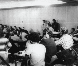 בתמונה השחור-לבן הזו של חדר ישיבות צפוף יושבים אנשים, חלקם אוזניות. כמה אנשים עומדים על הקיר, אחד מדבר לתוך מיקרופון. ציוד מגוון מציע מסיבת עיתונאים או הגדרת שידור על ידי חבר תרגומים.