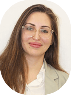 אישה עם שיער חום ארוך ומשקפיים מחייכת בעדינות. היא לובשת בלייזר בהיר מעל חולצה לבנה, על רקע לבן רגיל.