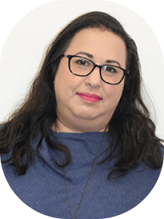 אישה עם שיער כהה ארוך המרכיבה משקפיים וחולצה כחולה מצטלמת מול רקע לבן רגיל. יש לה חיוך עדין והיא לובשת שפתון ורוד.