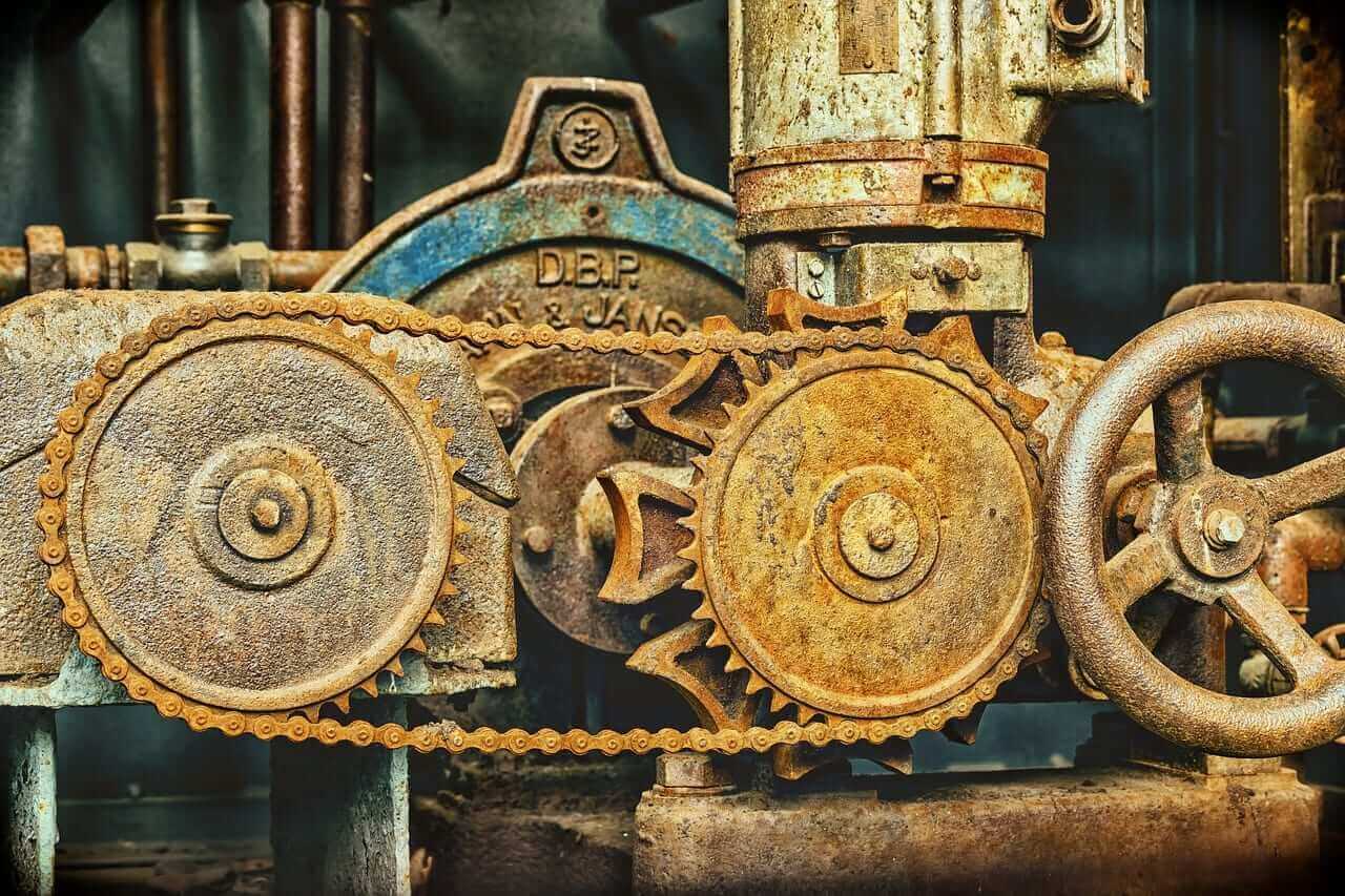 Close-up of rusty gears and a chain in an old industrial machine setup, featuring large circular metal components and a pipe extending vertically. The corroded surfaces evoke unanimous agreement on their vintage charm, creating an atmosphere where time seems to stand still.