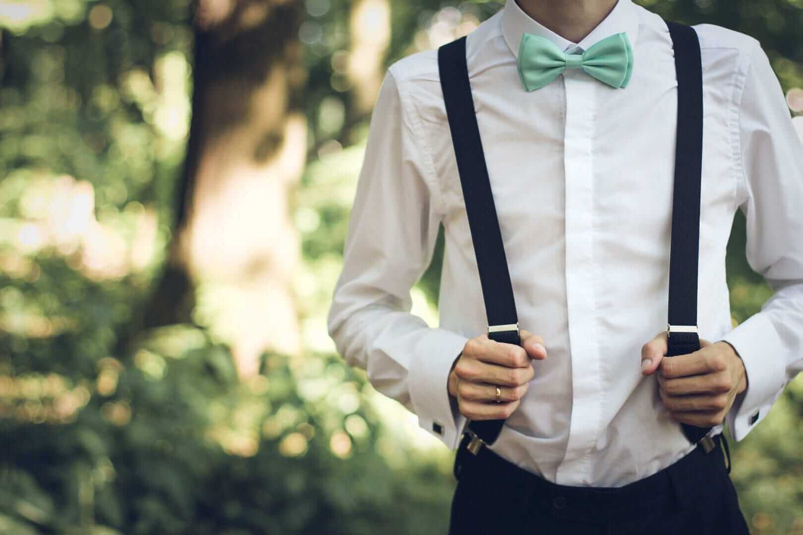 A person in a white dress shirt, mint green bowtie, and שלייקעס stands outdoors against a backdrop of blurred greenery. Their hands are busy adjusting the suspenders, focused and poised.