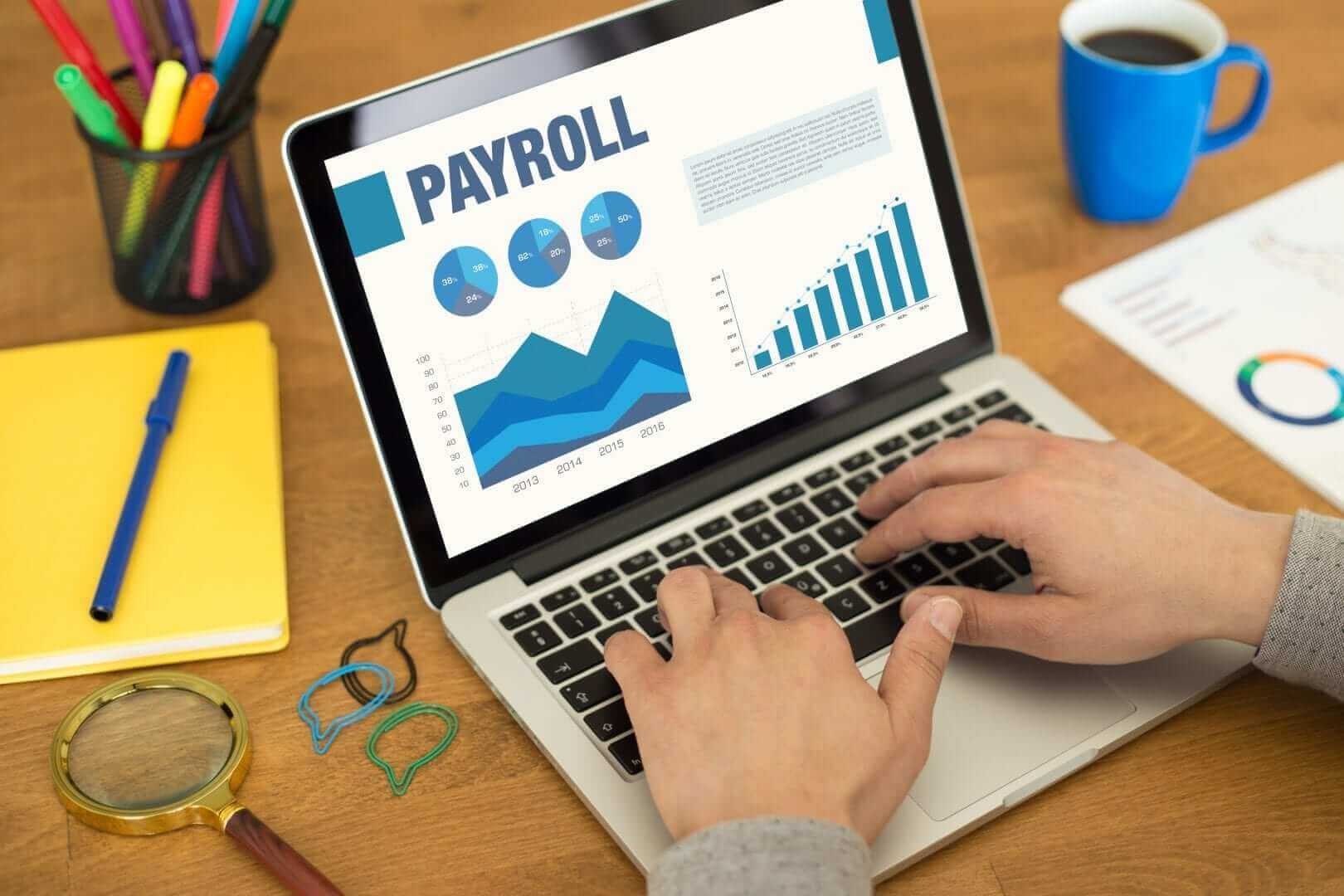 אדם מקליד על מחשב נייד ומציג לוח מחוונים של שכר עם תרשימים וגרפים, כנראה מהרהר מתי לתרגם תלוש. השולחן מאורגן עם זכוכית מגדלת, מהדקי נייר, פנקס צהוב, עט, סמארטפון וספל כחול.