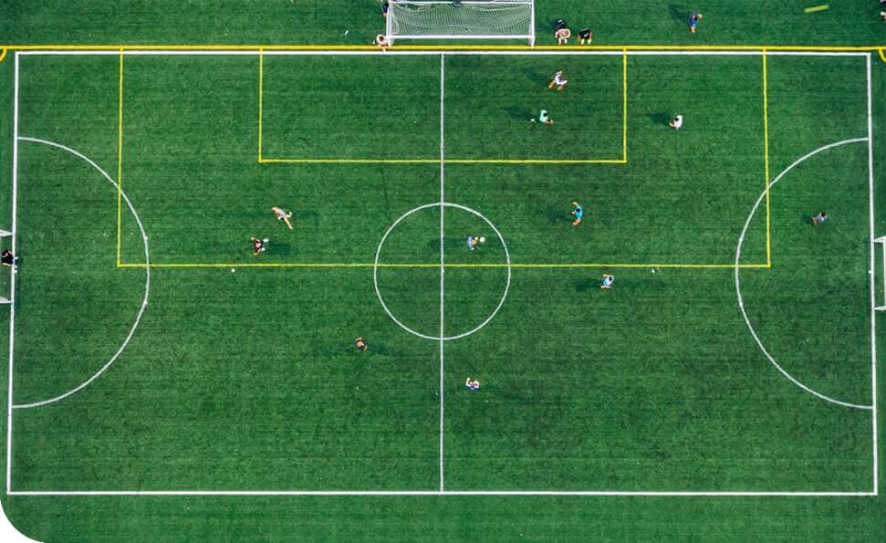 מבט אווירי של מגרש כדורגל, המגרש, עם שחקנים גלויים, מסומן בקווי גבול צהובים. במגרש הירוק הבהיר יש שחקנים מפוזרים בעמדות שונות, ועמוד השער נמצא בראש התמונה.