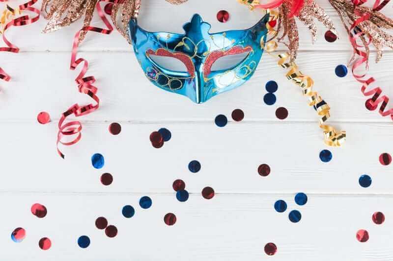 מסכת מסכות כחולה ואדומה עם פירוט זהב מונחת בין סטרימרים אדומים וזהובים, קונפטי מפוזר על משטח עץ לבן, מהדהדת את הרוח התוססת של עלון מרץ 2019 של חבר תרגומים.