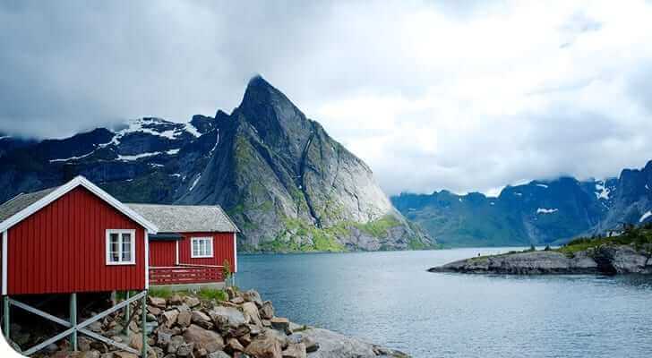 בקתה אדומה על כלונסאות יושבת על קו חוף סלעי מתחת לשמים מעוננים, המזכירה סצנה היישר מניוזלטר. פיורד כחול ורגוע נמתח לעבר הרים דרמטיים ותלולים ברקע, עטופים חלקית בערפל, המעורר את הפיתוי השלו שנלכד במאי 2020.