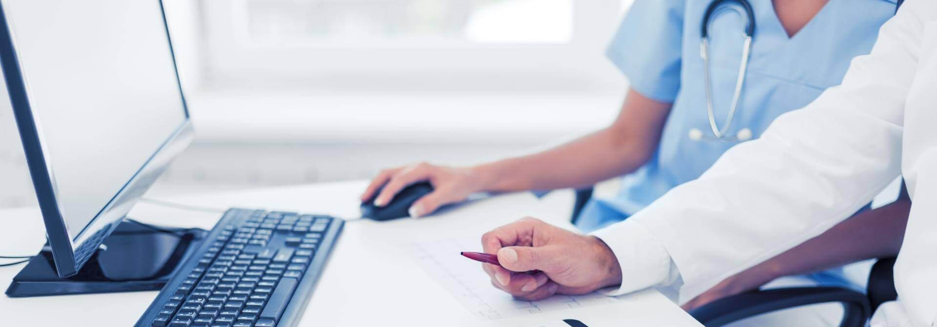 שני אנשי מקצוע בתחום הבריאות יושבים ליד שולחן. האחד, במעיל לבן, מצביע על מסמך בעט אדום הקשור לתרגום נוטריוני, ואילו השני, בקרצוף תכלת, משתמש בעכבר מחשב. מקלדת ומסך גלויים על השולחן.