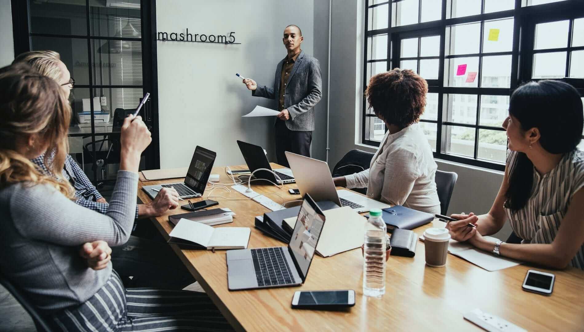 קבוצה של שישה אנשים מכונסת בחדר ישיבות. אדם אחד עומד בחזית עם טוש, מציג בפני חמישה יושבים המשתמשים במחשבים ניידים לתמלול ישיבות ורושם הערות. חלון גדול מכניס אור טבעי, ועל הקיר כתוב mashroom5.