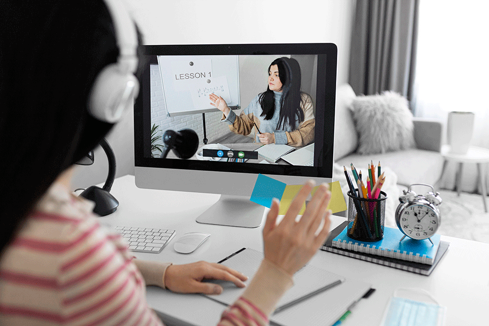 אדם חובש אוזניות עוסק בשיעור מקוון על מסך מחשב. התצוגה חושפת מורה אוחזת במחברת בזמן שהאדם מנופף. מוקף במחברת, שעון וכלי כתיבה, סצנה זו לוכדת את המהות של חינוך גלובלי בלמידה מקוונת מודרנית.