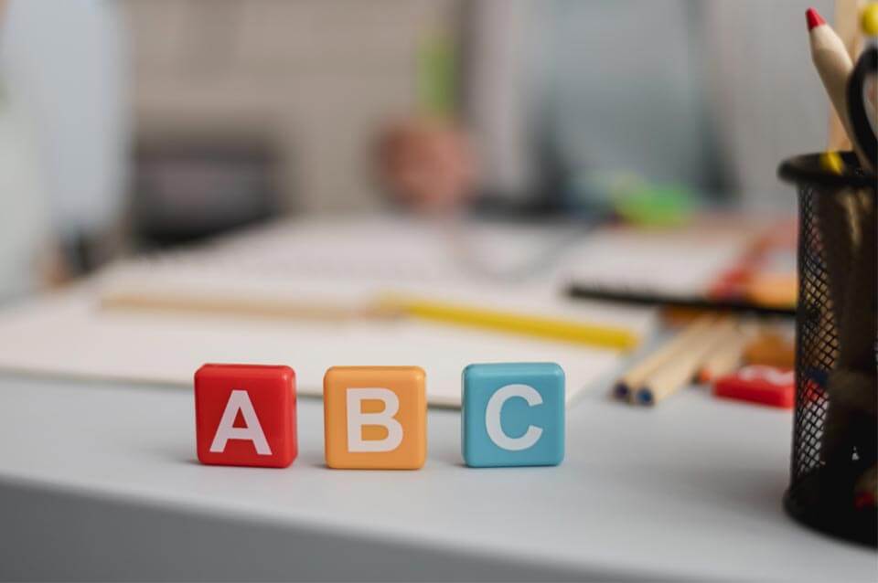 שלושה בלוקים צבעוניים המציגים את האותיות A, B ו-C מסודרים על שולחן. ברקע חפצים מטושטשים, ביניהם מחזיק עפרונות, מחברת ופתקים על עברית לאנגלית לעבודות תרגום.