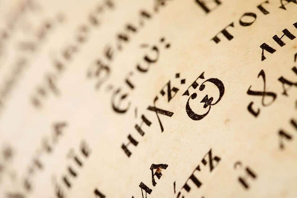 תקריב של עמוד המציג טקסט ישן ומעוטר בגופן סריף. הנייר המיושן והצהוב חושף תרגום שפות בין הדמויות המובהקות במרכז, ומרמז על משמעות היסטורית או דתית לאורך האבולוציה של הכתיבה לאורך השנים.