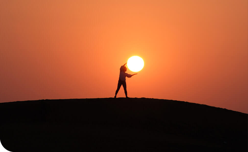 צללית של אדם עומד על גבעה בשעת השקיעה, נראה מחזיק את השמש בידיו. השמיים הם שיפוע של כתום ואדום, היוצרים סצנה דרמטית ושלווה.