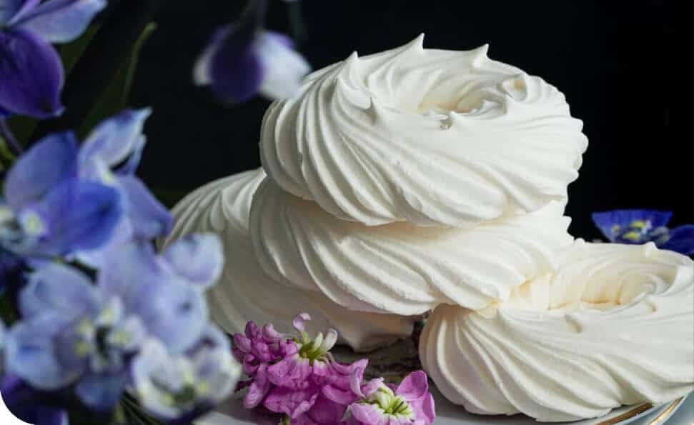 שלוש מערבולות מרנג לבנות מוערמות על משטח, עטפות בפרחים סגולים וורודים. הרקע הכהה משפר את הניגודיות של המרנג והפריחה, ויוצר פינוק ויזואלי שהוא באמת ביס מעולם אחר.