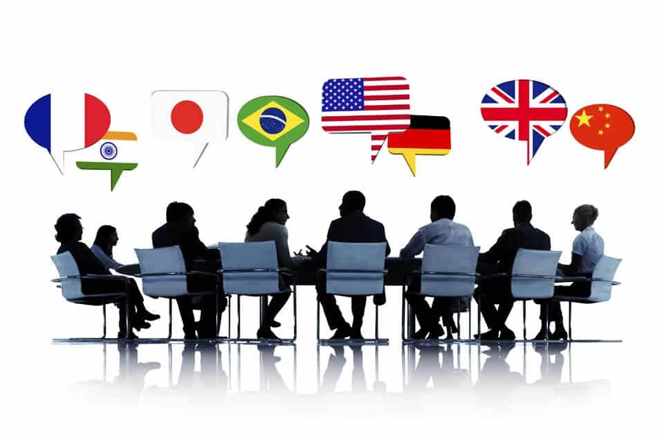צלליות של אנשים יושבים ליד שולחן, עם פגישה עם בועות דיבור מעליהם המציגות דגלים מצרפת, הודו, יפן, ברזיל, ארהב, גרמניה, בריטניה וסין, מה שמציע לוקליזציה או שיתוף פעולה בינלאומי בעולם רב לשוני.