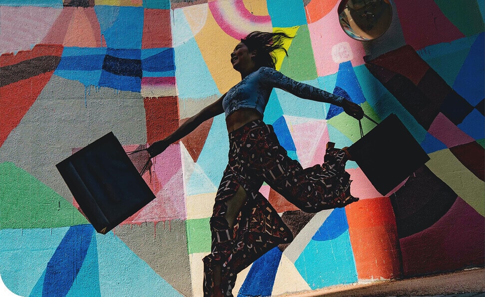 אדם מזנק בשמחה מול ציור קיר צבעוני, אוחז בשקיות שופינג. הרקע החי כולל דפוסים גיאומטריים מופשטים, והאדם מטיל צל מובהק, ומדגיש את התנועה הדינמית שלו.