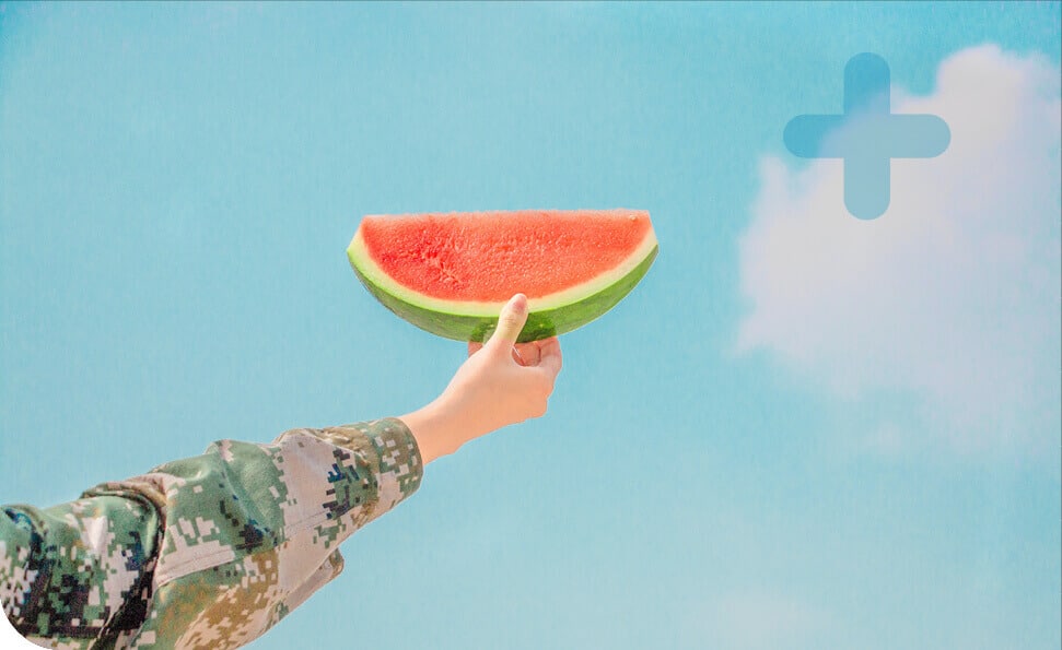 יד בשרוול בדוגמת הסוואה מחזיקה פרוסת אבטיח על רקע השמיים הכחולים והצלולים, כאשר ענן קלוש וצורת סימן פלוס מדגישים את הסצנה. אחרי הכל, עיקר הבריאות.