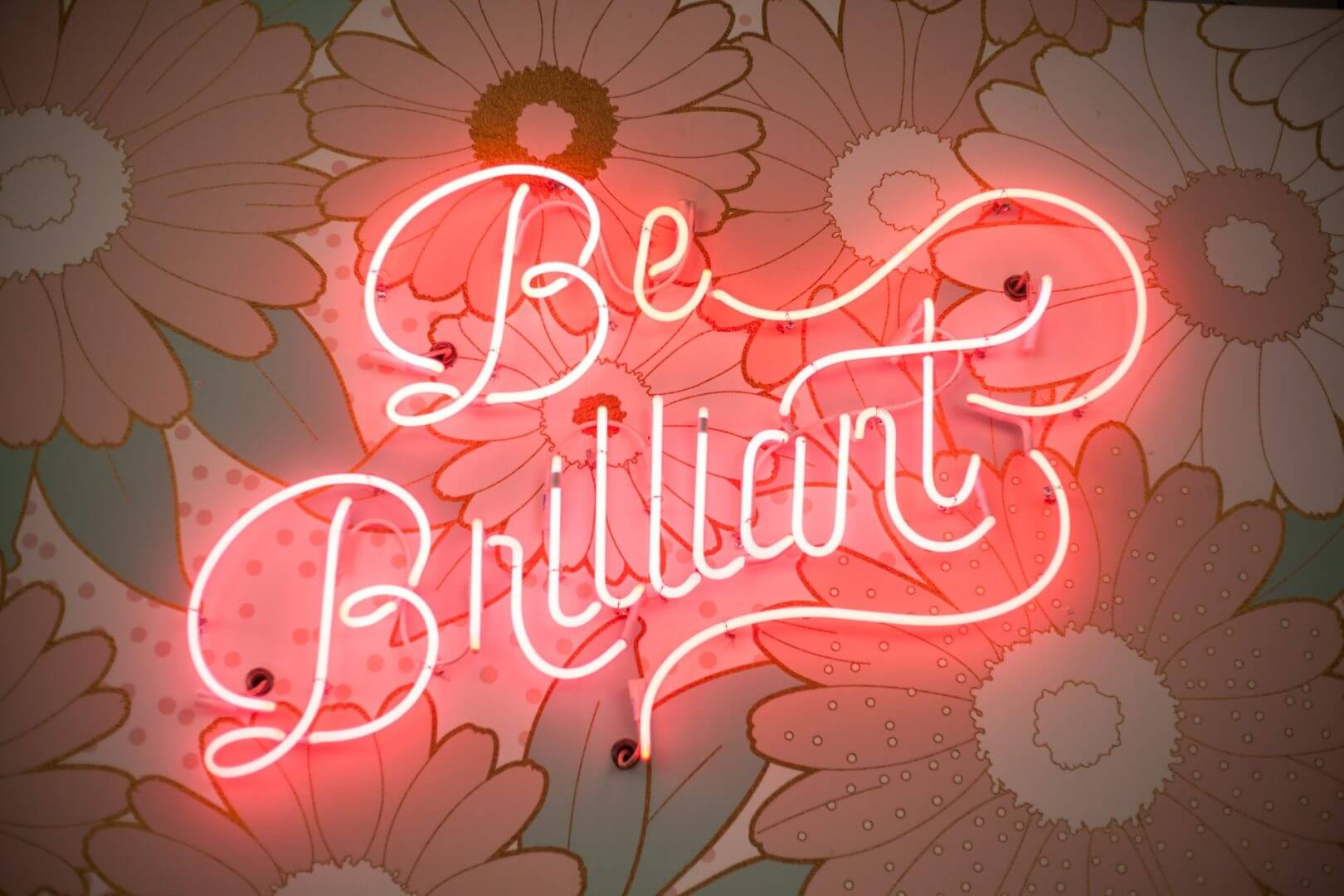 שלט ניאון ורוד כתוב Be Brilliant על רקע טפט פרחוני עם פרחים דמויי דייזי בצבעי פסטל רכים, המסמלים את הפתרונות הבהירים שנמצאים באתגרי התרגום.