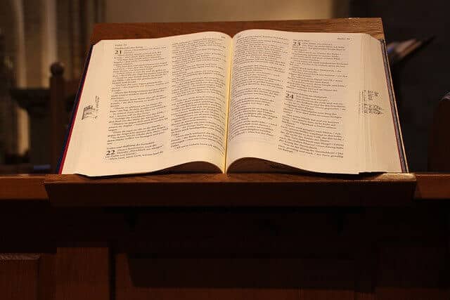 תנך פתוח מונח על דוכן עץ בחדר אפלולי. הדפים, המזכירים את הספרים המתורגמים ביותר, מציגים טקסט מודפס מקרוב, והתפאורה מציעה סביבה של כנסייה או קפלה.