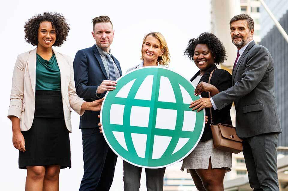 קבוצה מגוונת של חמישה אנשים לבושים במקצועיות עומדת בחוץ, מחייכים ואוחזים בסמל גלובוס ירוק גדול, המגלם רוח של עסק גלובלי. הרקע מציג אלמנטים מטושטשים של נוף עירוני, המדגיש את הקשר העירוני שלהם ומתמקד בלוקיזציה בפרקטיקות העסקיות.