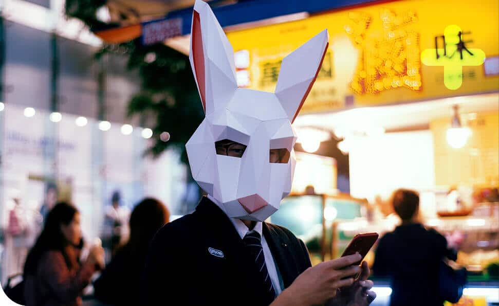 בסצנת העיר הסואנת, אדם בחליפה ומסיכת ארנב גיאומטרית בודק את הסמארטפון שלו, בין דמויות מטושטשות ושילוט תוסס. התפאורה התוססת מרמזת על מסיבת תחפושות יוצאת דופן שמתגלה בסמוך.