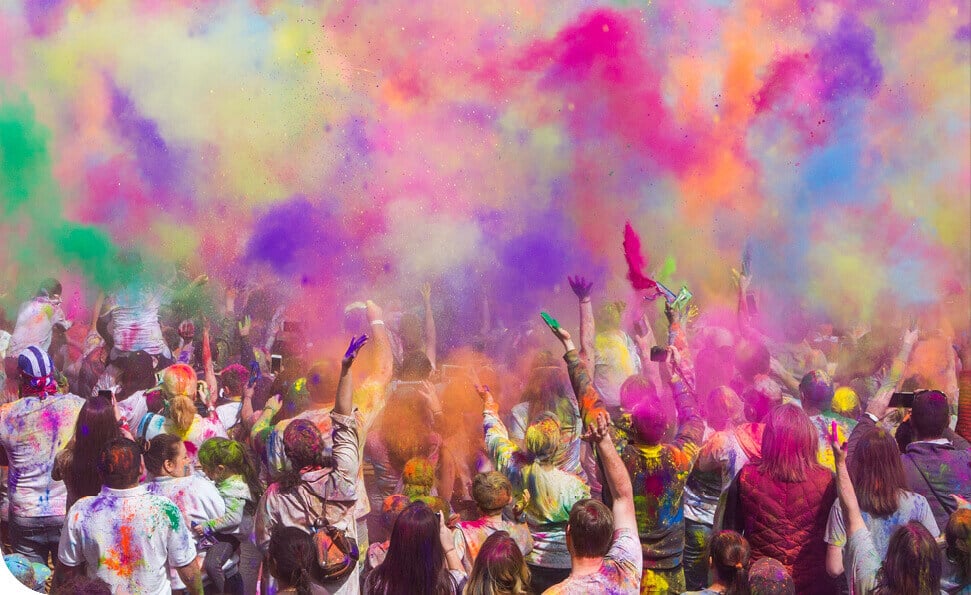 קהל גדול חוגג עם אבקות צבעוניות עזות שנזרקות לאוויר, ויוצרות ענן של גוונים ורודים, כתומים, סגולים וירוקים. המשתתפים מכוסים בצבעים, מתענגים על אווירת הפסטיבל החיצונית התוססת כשהדים של עוד חוזר הניגון ממלאים את האוויר.
