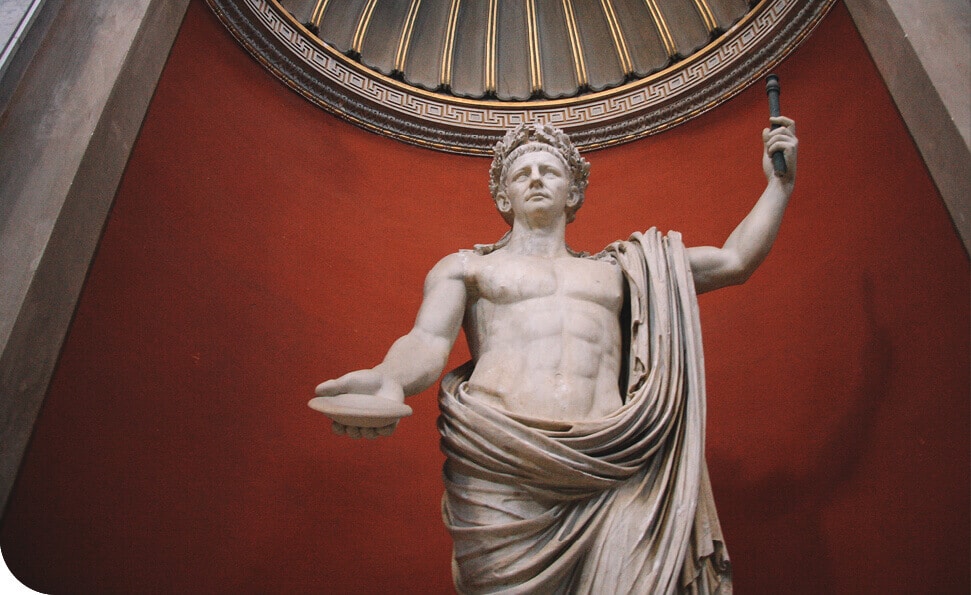 פסל של דמות גברית שרירית קובע שיאים בנוכחותו השלטת, אוחזת שרביט ביד אחת ובשנייה צלחת, עטופה באלגנטיות בבד. הוא עומד על קיר אדום המעוטר באלמנט דקורטיבי בצורת חצי עיגול מעל.