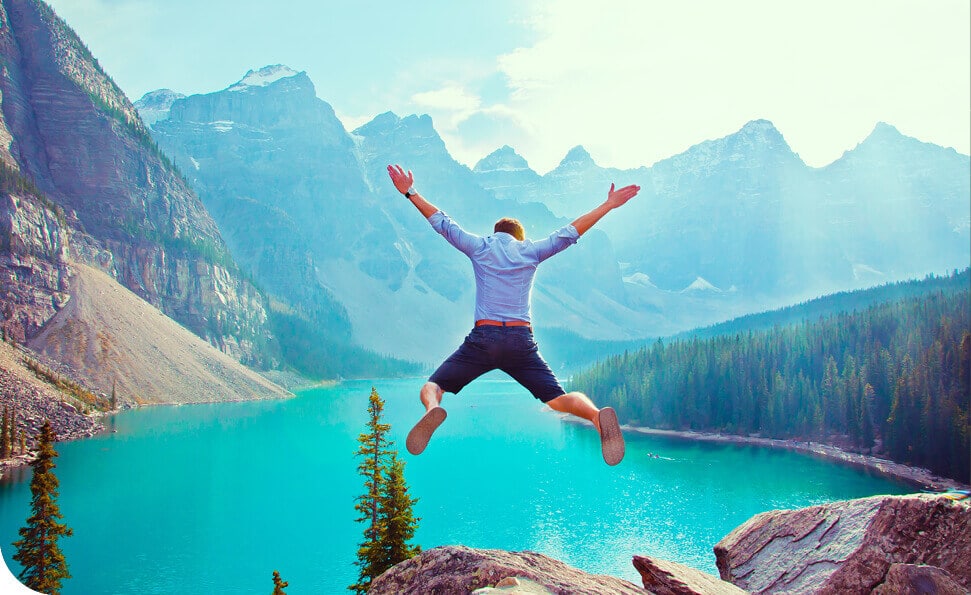 אדם בחולצה כחולה ומכנסיים קצרים כהים קופץ בשמחה על סלע המשקיף על אגם בצבע טורקיז, מוקף בעצי אורן ובהרים מחוספסים מתחת לשמים כחולים צלולים. כאילו חוגגים את תחילת השנה, אור השמש מדגיש את הפסגות ואת הצבע העז של המים, ויוצר סצנה ראויה להערצה.