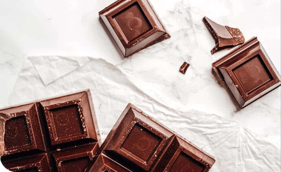 תקריב של חתיכות שוקולד מריר שבורות על נייר פרגמנט מקומט, מזכיר בונבוניירה יוקרתית. השוקולד מתהדר במרקם חלק עם ריבועים עמוקים ומבריק, המפוזרים באלגנטיות על פני הנייר. הרקע כולל משטח בהיר ומשיש.