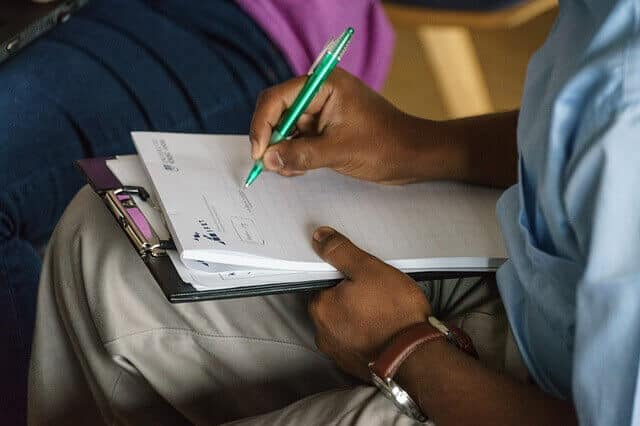 אדם כותב בקפידה בעט ירוק על לוח כתיבה מלא בניירות, יושב ברגליים משוכלות. הפוקוס הוא על היד והניירות שלהם, בעוד הרקע חושף חלק של אדם אחר בגינס. זה כאילו הם בוחרים בין תמלול אוטומטי או תמלול אנושי להערות שלהם.