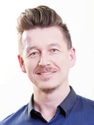 גבר עם שיער חום מעוצב ושפם מחייך למצלמה. הוא לובש חולצה מכופתרת כחולה כהה. הרקע לבן רגיל.