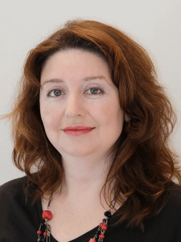 אדם עם שיער חום גלי ולובש עליונית שחורה ושרשרת צבעונית מחייך קלות. הרקע אפור רגיל.