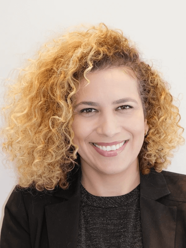 אדם עם שיער בלונדיני מתולתל מחייך למצלמה. הם לובשים בלייזר שחור מעל חלק עליון כהה. הרקע לבן רגיל.