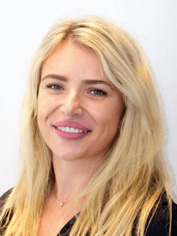 אדם חייכן עם שיער בלונדיני ארוך, עונד חלק עליון כהה ושרשרת דקה, מול רקע לבן רגיל.