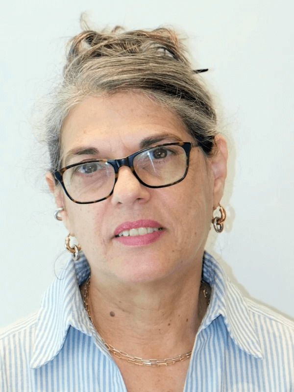 אישה עם משקפיים ושיער מאפיר קשורה, לובשת חולצת פסים כחול-לבן ועגילי חישוק זהב, עומדת על רקע בהיר, מביטה במצלמה בהבעה ניטרלית.