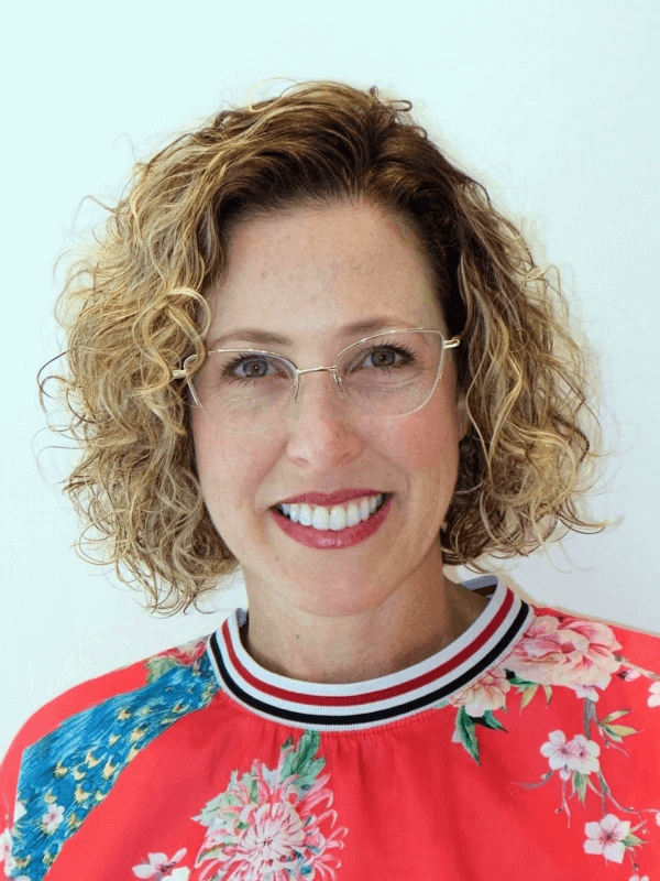 אדם עם שיער בלונדיני מתולתל ומשקפיים מחייך למצלמה. הם לובשים חולצה בהירה בדוגמת פרחים עם צווארון פסים. הרקע לבן רגיל.