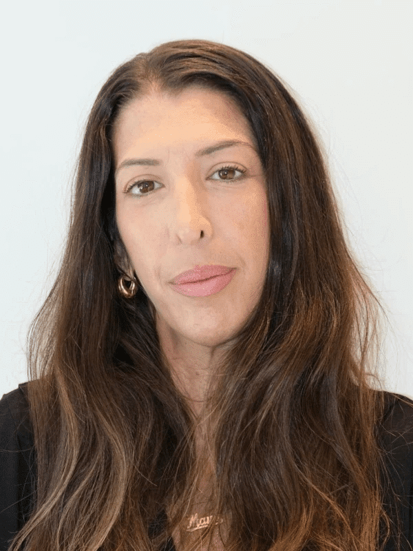 אדם עם שיער חום ארוך, עונד חלק עליון שחור ועגילי חישוק זהב, על רקע בהיר.