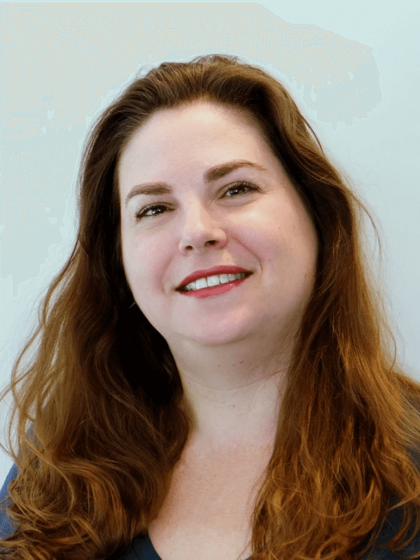 אדם עם שיער חום ארוך וגלי מחייך למצלמה. הם לובשים חלק עליון כחול, והרקע הוא בצבע בהיר ופשוט.