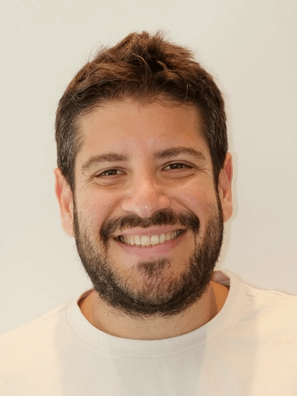 אדם עם שיער כהה קצר וזקן מחייך למצלמה. הם לובשים חולצה בהירה על רקע רגיל.