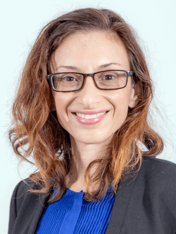 אדם חייכן עם שיער חום גלי ומשקפיים, לובש חלק עליון כחול ובלייזר שחור, עומד על רקע רגיל.