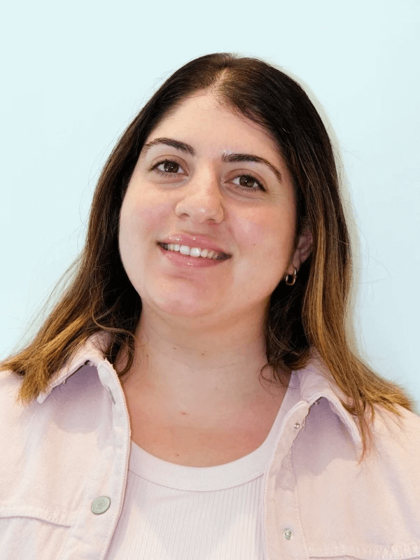 אדם חייכן עם שיער חום ארוך, לבוש בחולצה ורודה בהירה וחולצה לבנה, על רקע תכלת רגיל.