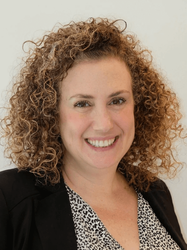 אישה עם שיער מתולתל מחייכת למצלמה. היא לובשת בלייזר שחור מעל חולצה מעוצבת ועומדת על רקע לבן רגיל.