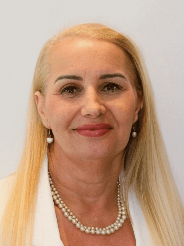 אישה עם שיער בלונדיני בהיר ארוך מחייכת ברכות. היא עונדת חלק עליון לבן, שרשרת פנינים ועגילי פנינים תואמים. הרקע בצבע בהיר רגיל.