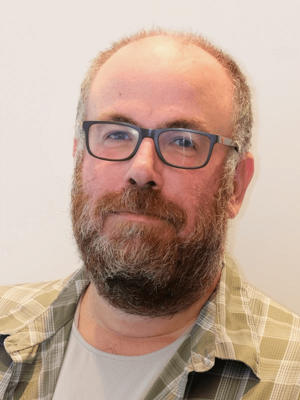 גבר מזוקן עם משקפיים, לובש חולצה משובצת מעל חולצת טריקו בהירה, עומד על רקע בהיר ופשוט.